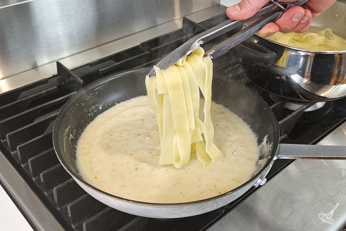 Someone placing cooked pasta in a frying pan with alfredo sauce. 