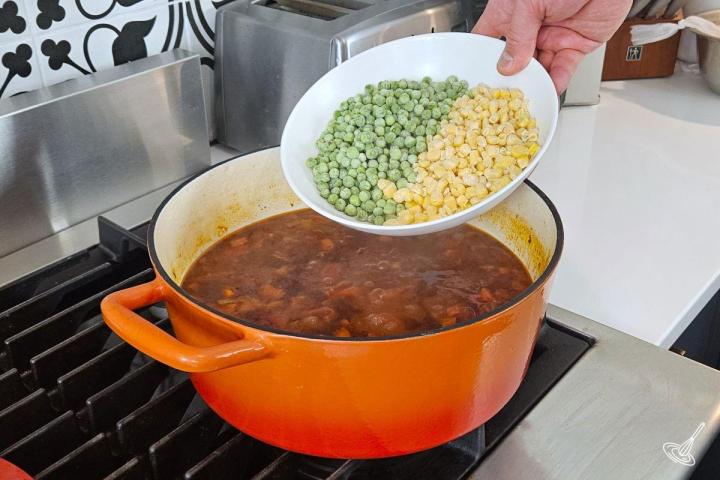 Someone placing frozen peas and corn kernels in a Dutch oven of soup. 