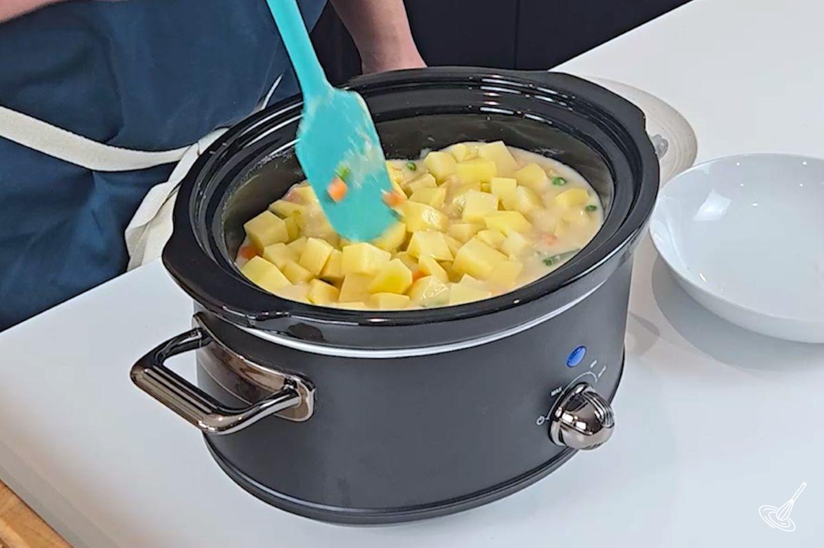 Someone stirring in cubed potatoes into a slow cooker with a cream sauce and vegetables.