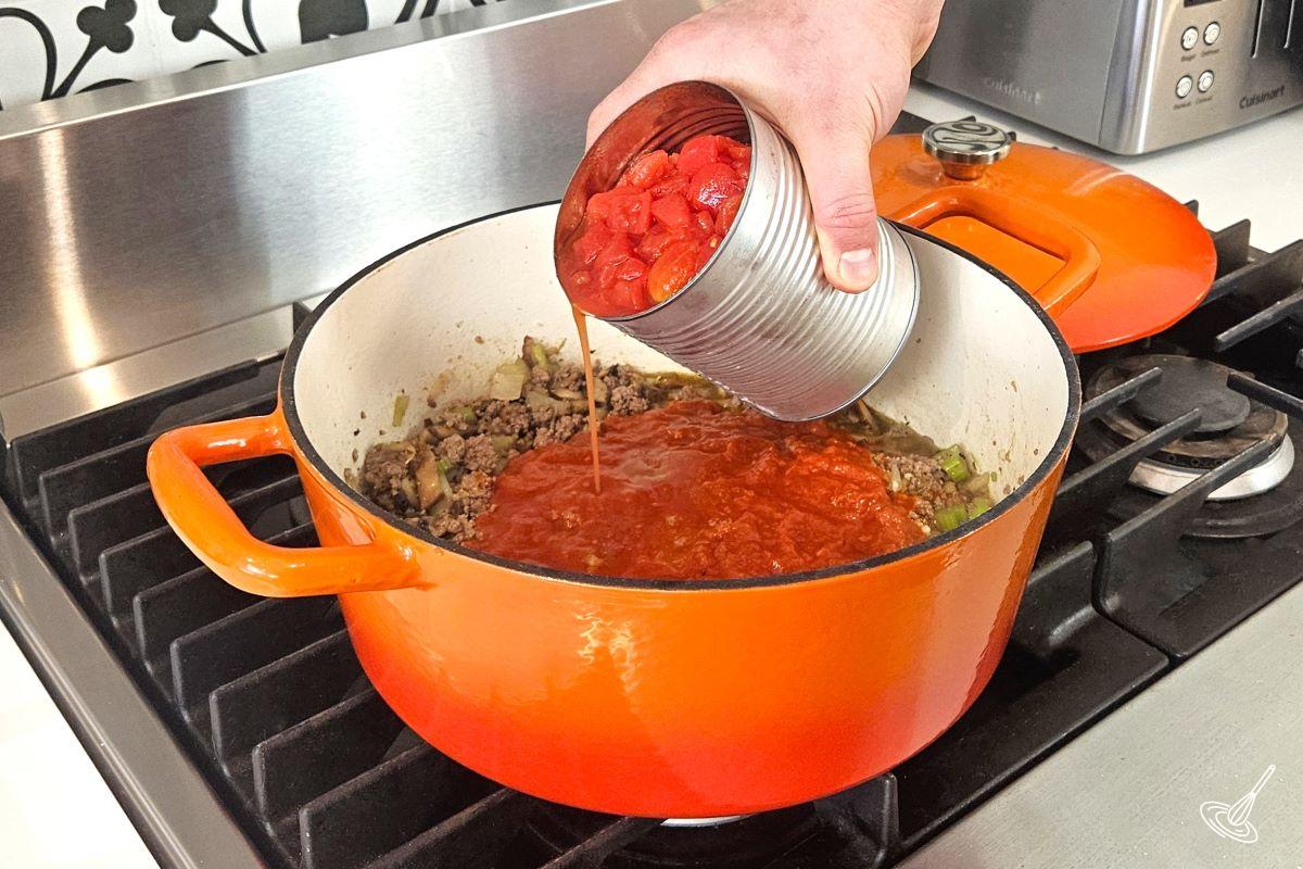 Someone pouring a can of tomato cubes in a Dutch oven with cooked ground beef and vegetables.