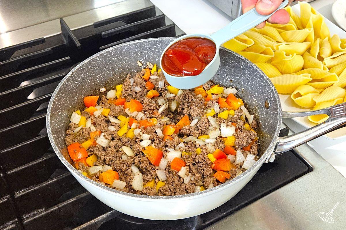 Someone adding a measuring cup of ketchup to a large skillet of cooked ground beef and veggies.