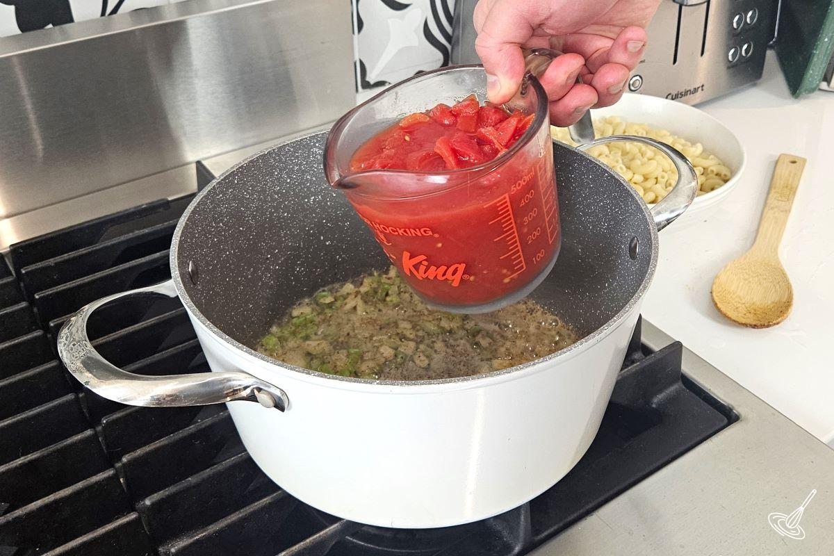 Someone adding canned tomatoes to a large pot with veggies and spices.
