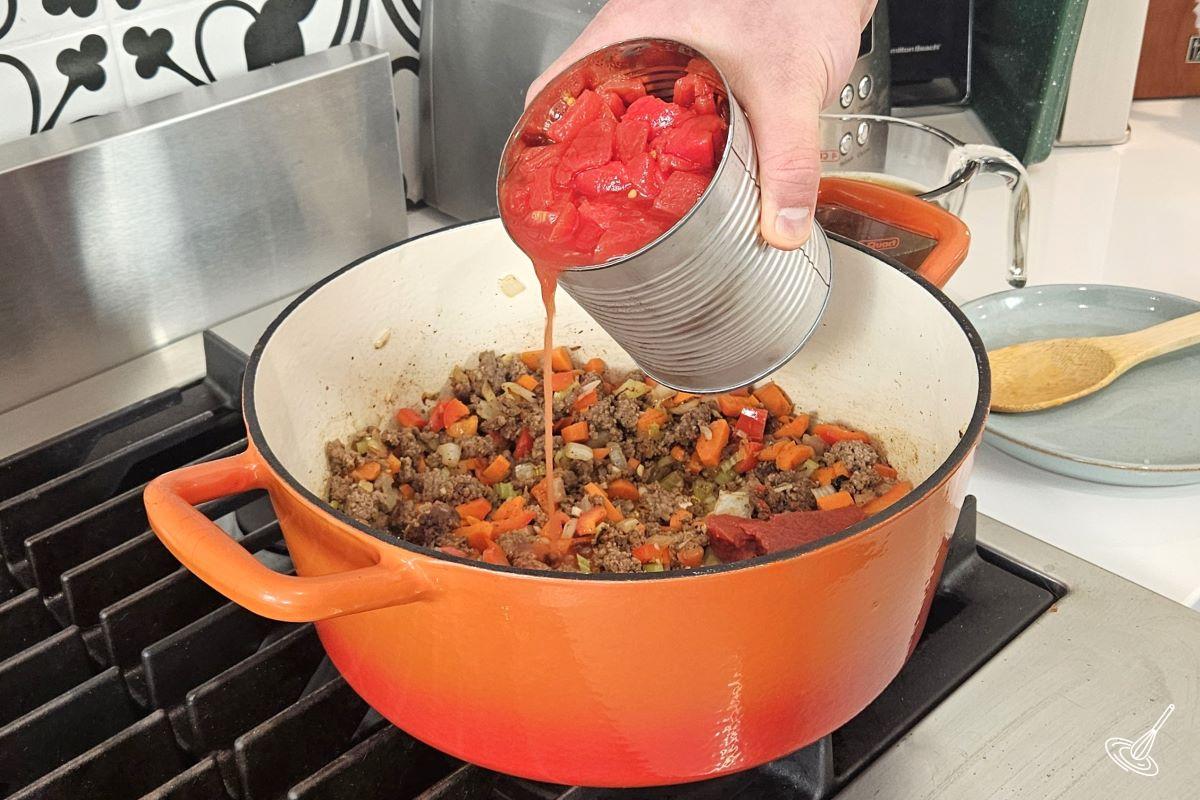 Someone pouring a can of tomatoes into a large pot with ground beef and tomatoes.