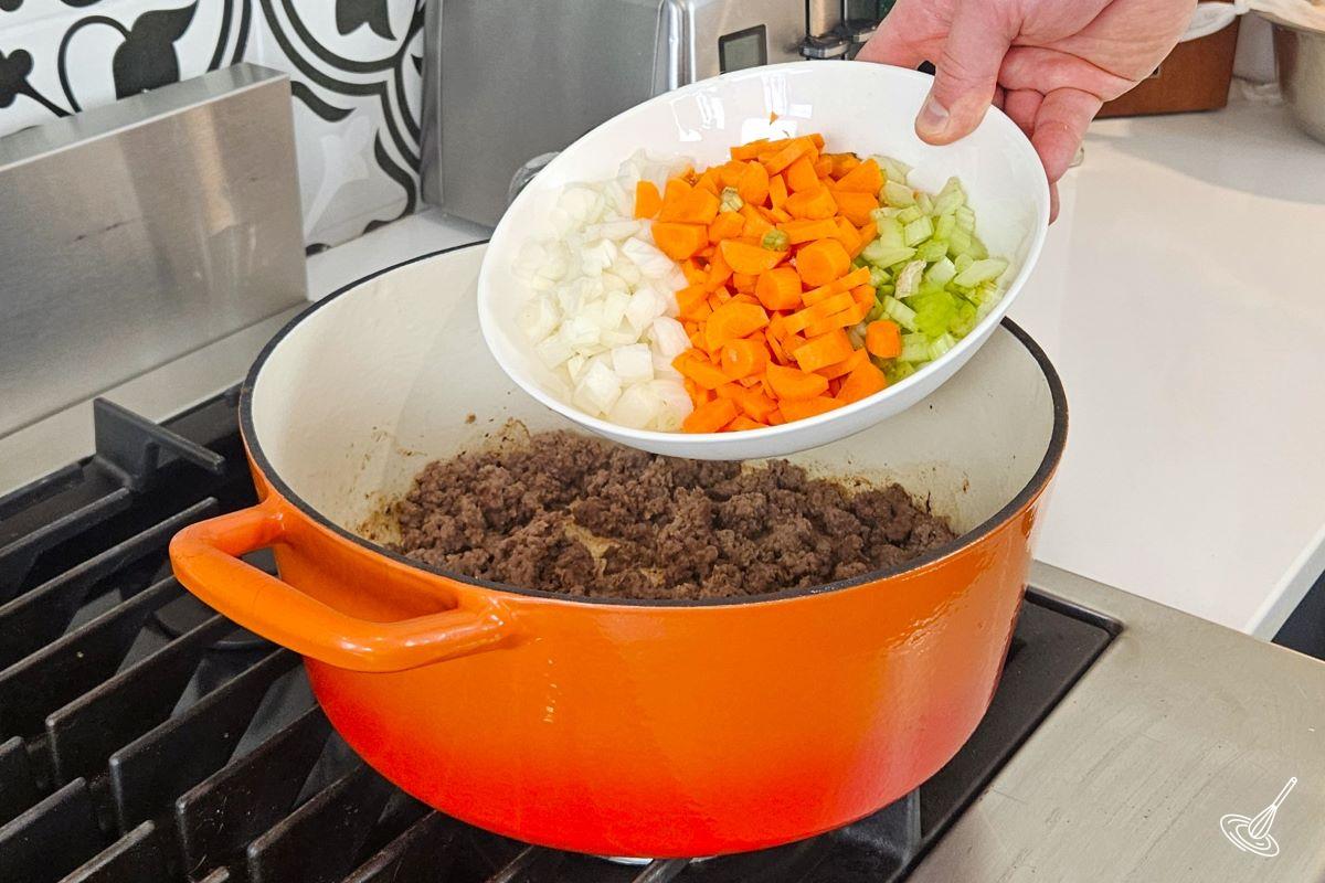 Someone placing chopped vegetables into a large pot on the stove with ground beef.