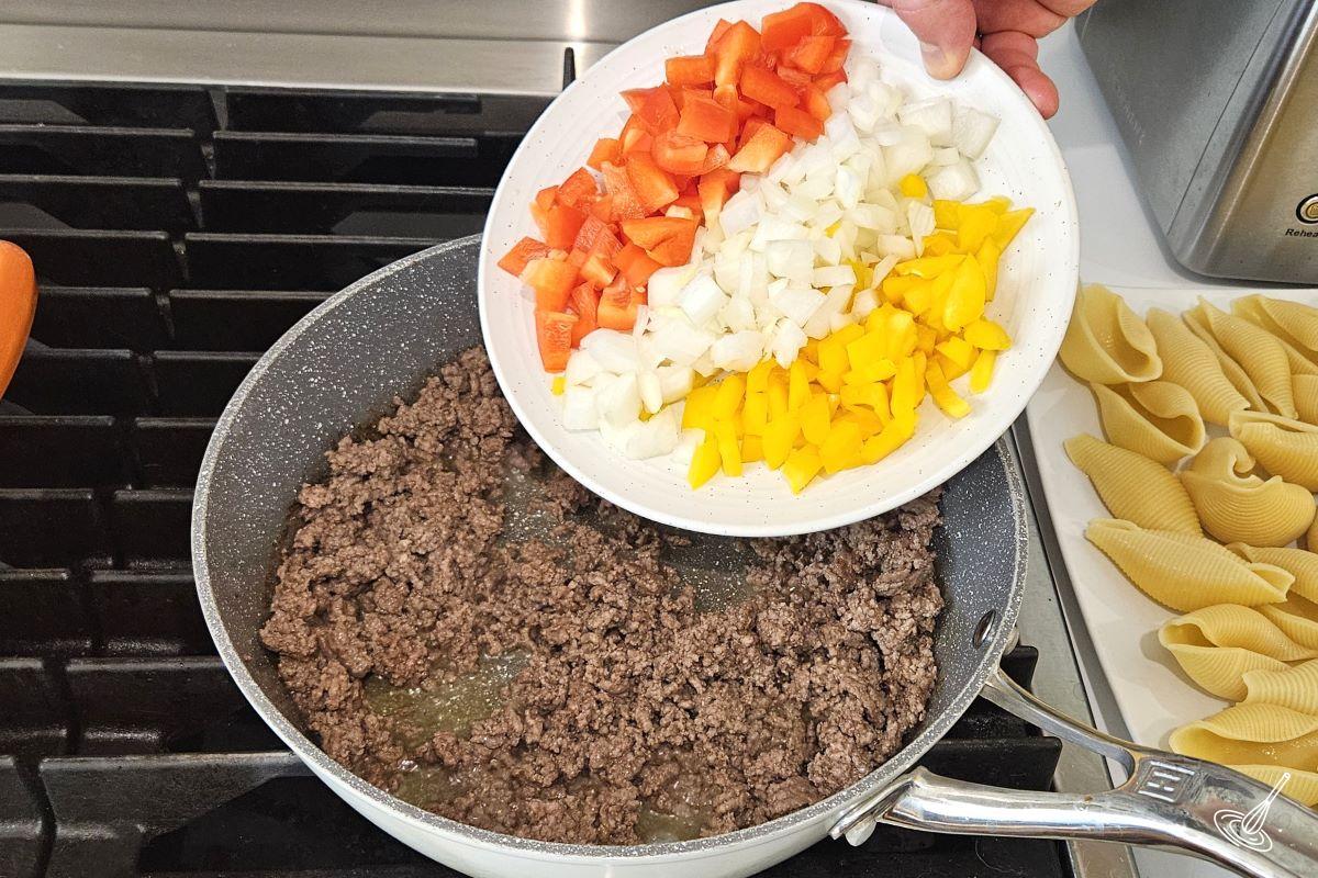 Someone adding chopped peppers and onions to the skillet of ground beef. 