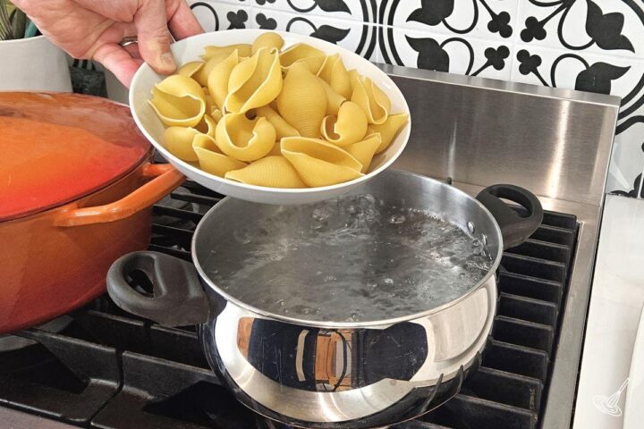 Someone placing a bowl of pasta shells into a large pot of boiling water.