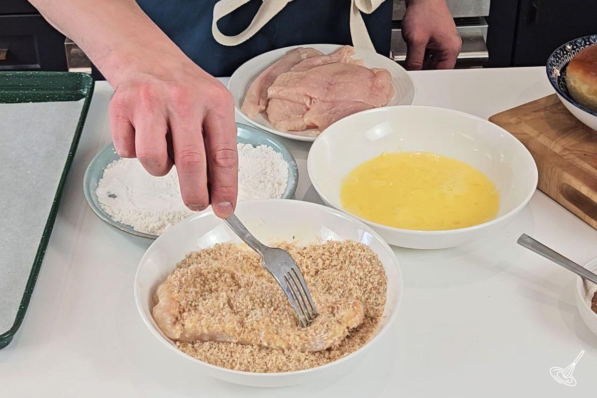 Someone coating a flattened chicken breast in egg, flour, and crispy coating. 