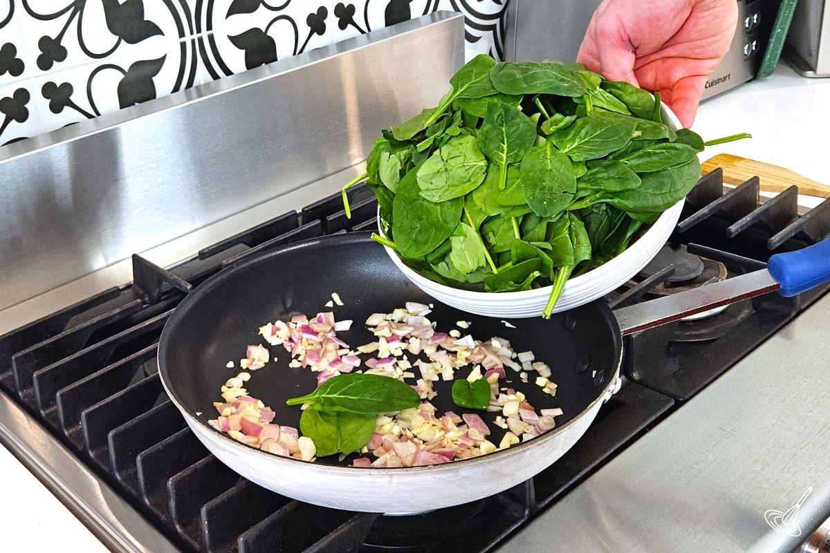 Someone placing a bowl of fresh spinach in a frying pan with onions and garlic. 