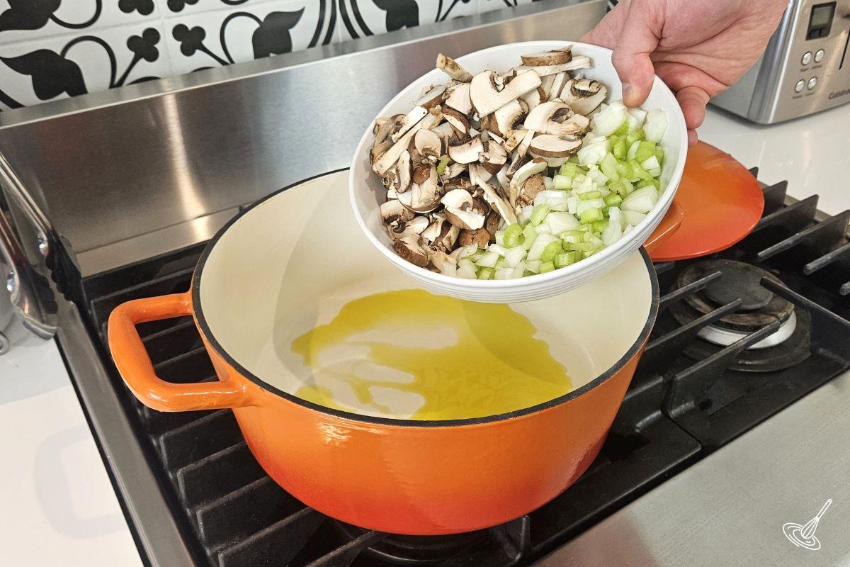 Someone placing chopped vegetables in a large Dutch oven with olive oil.