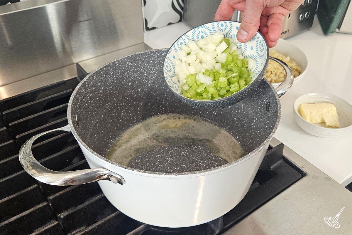 Someone adding chopped celery and onion to a large pot with melted butter.