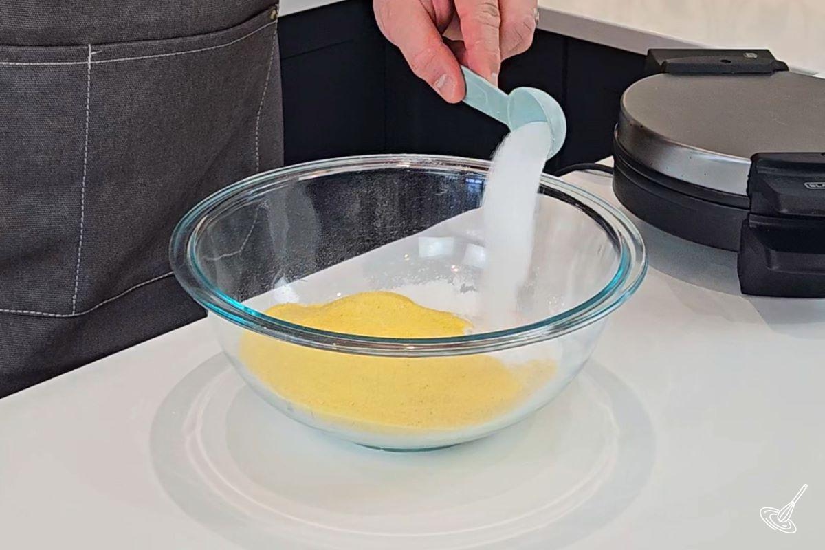 Someone adding baking powder to a large bowl with cornmeal and flour.