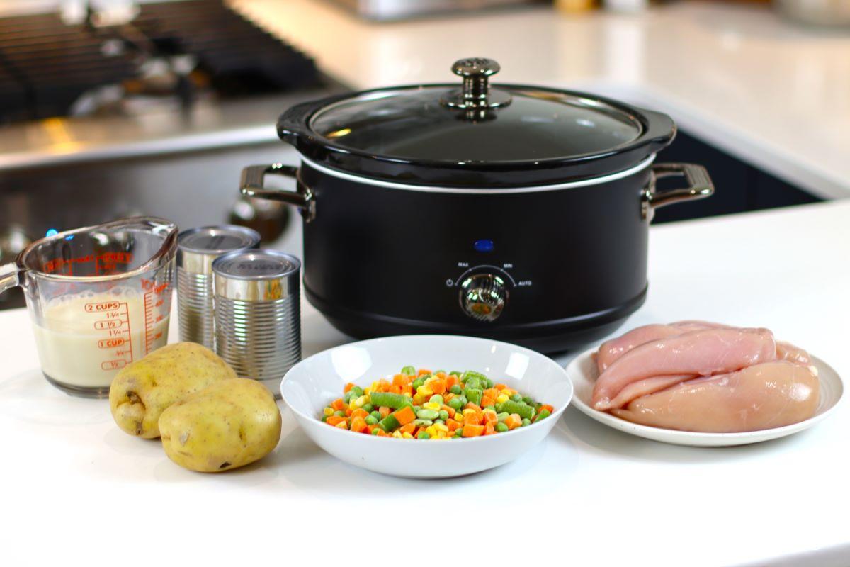 A slow cooker on the kitchen counter with a plate of chicken breasts and a bowl of frozen vegetables. 