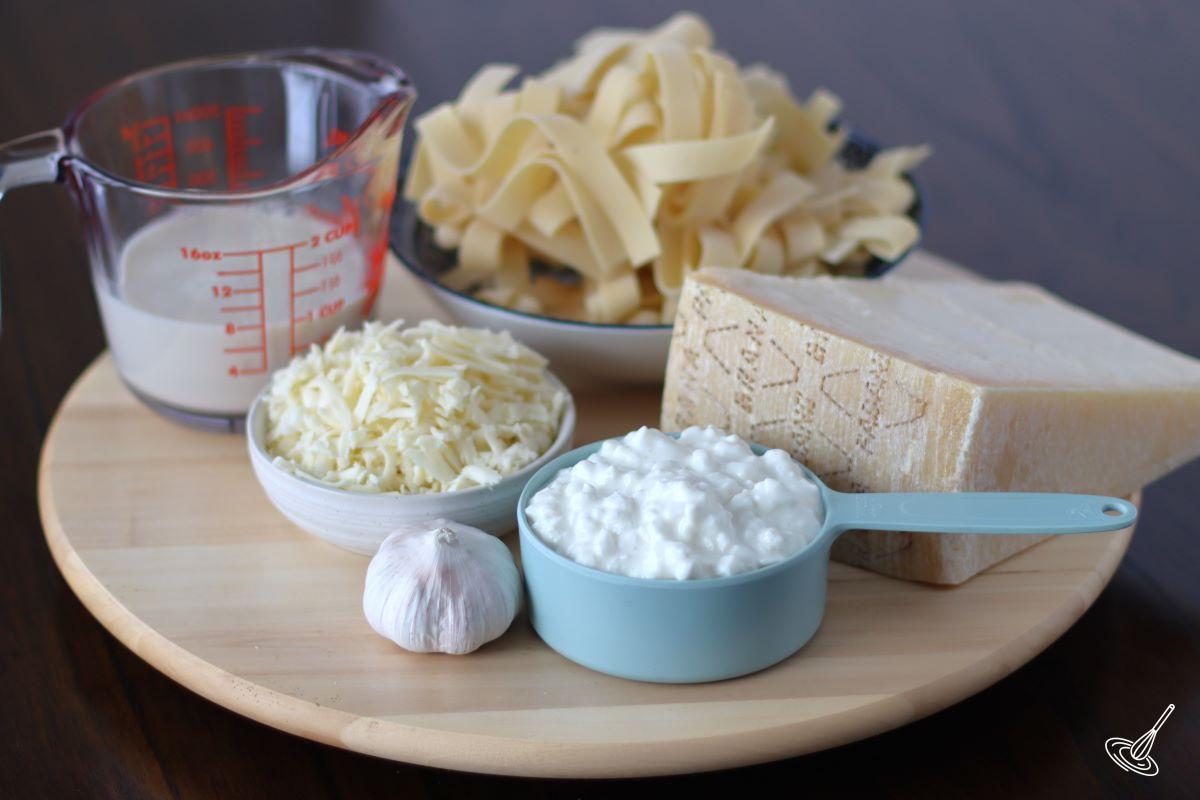 Ingredients on a wooden tray, including cottage cheese, grated parmesan, milk, and raw pasta.