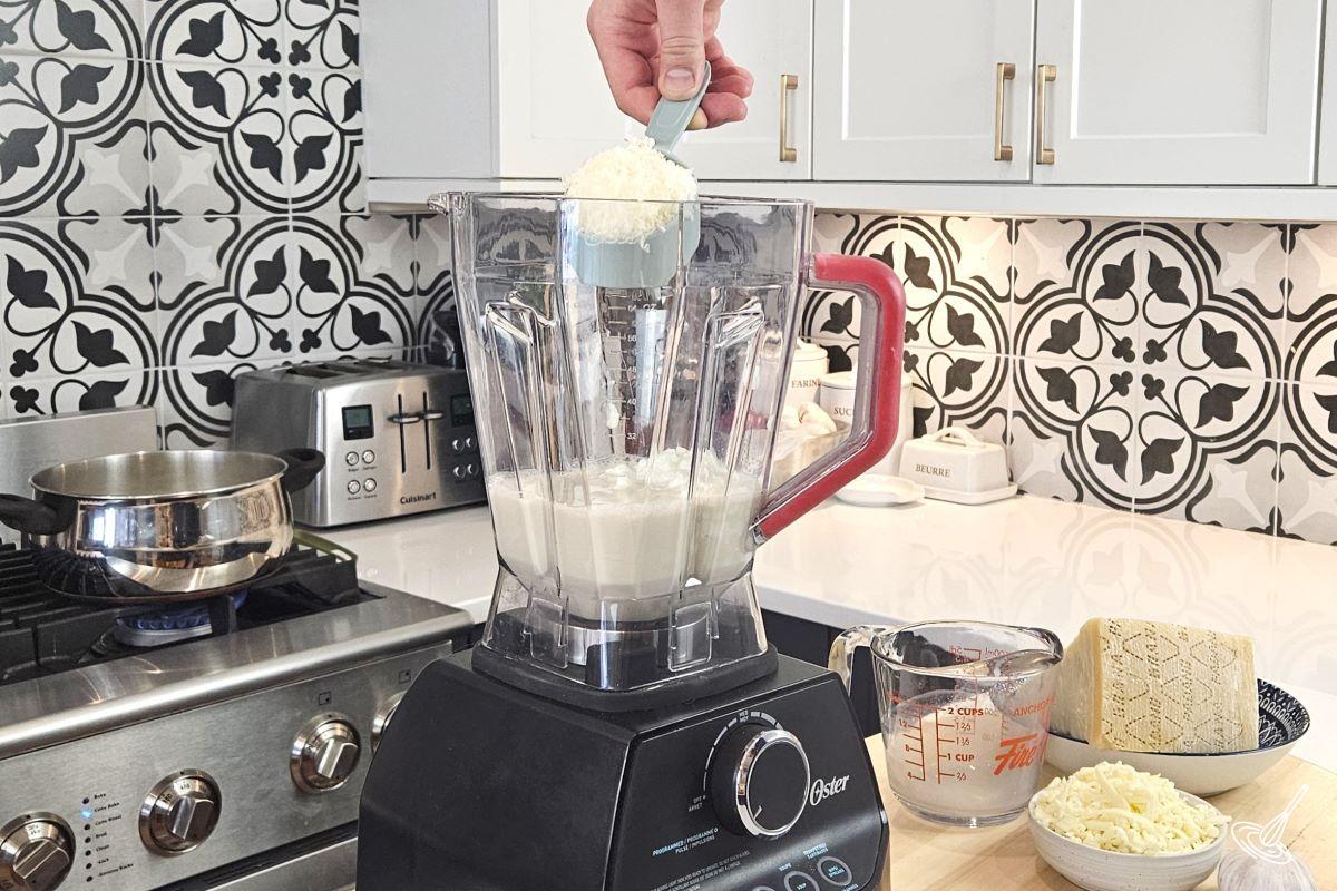Someone placing grated cheese in a blender with milk and cottage cheese.
