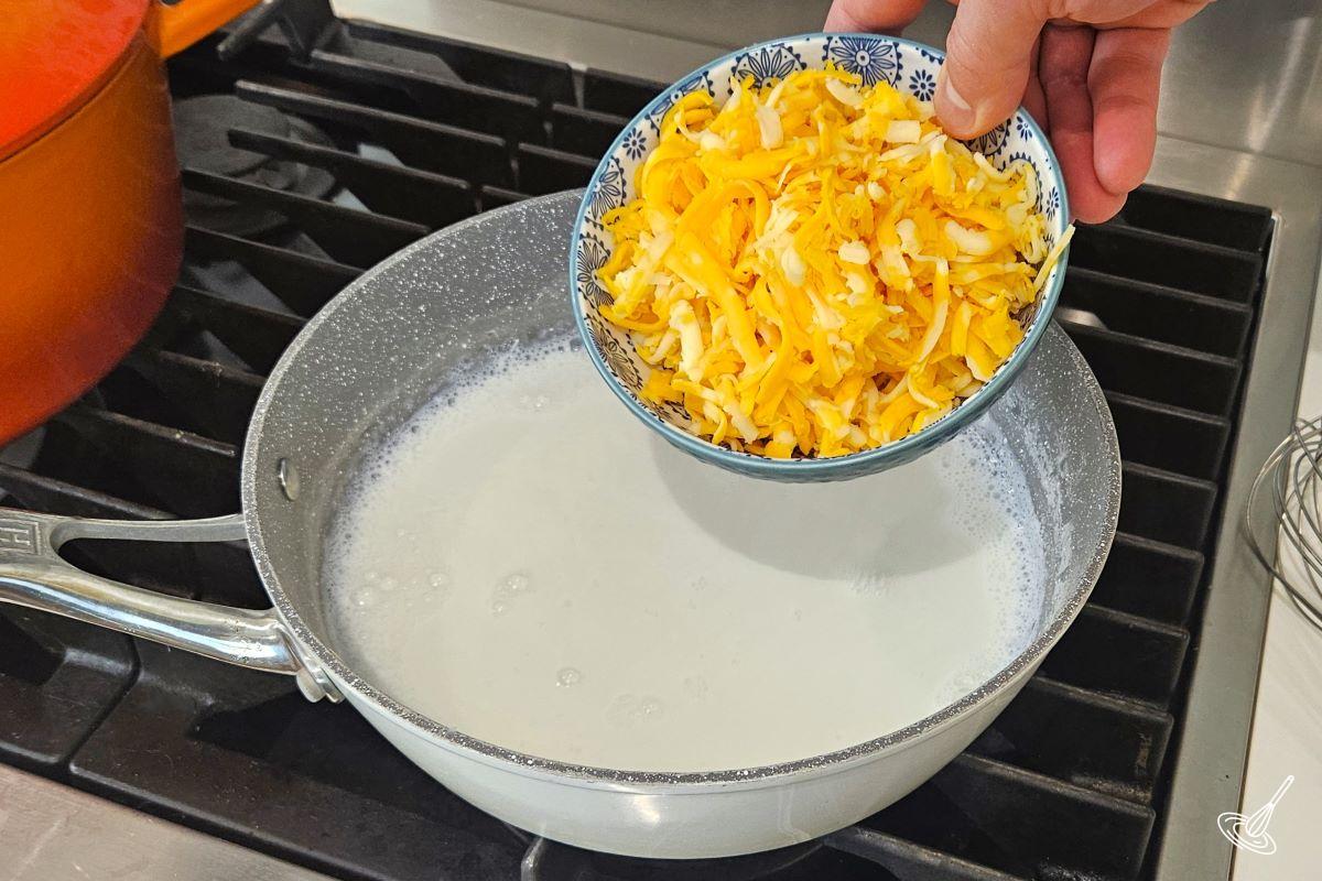 Someone placing shredded cheese in a saucepan with milk. 