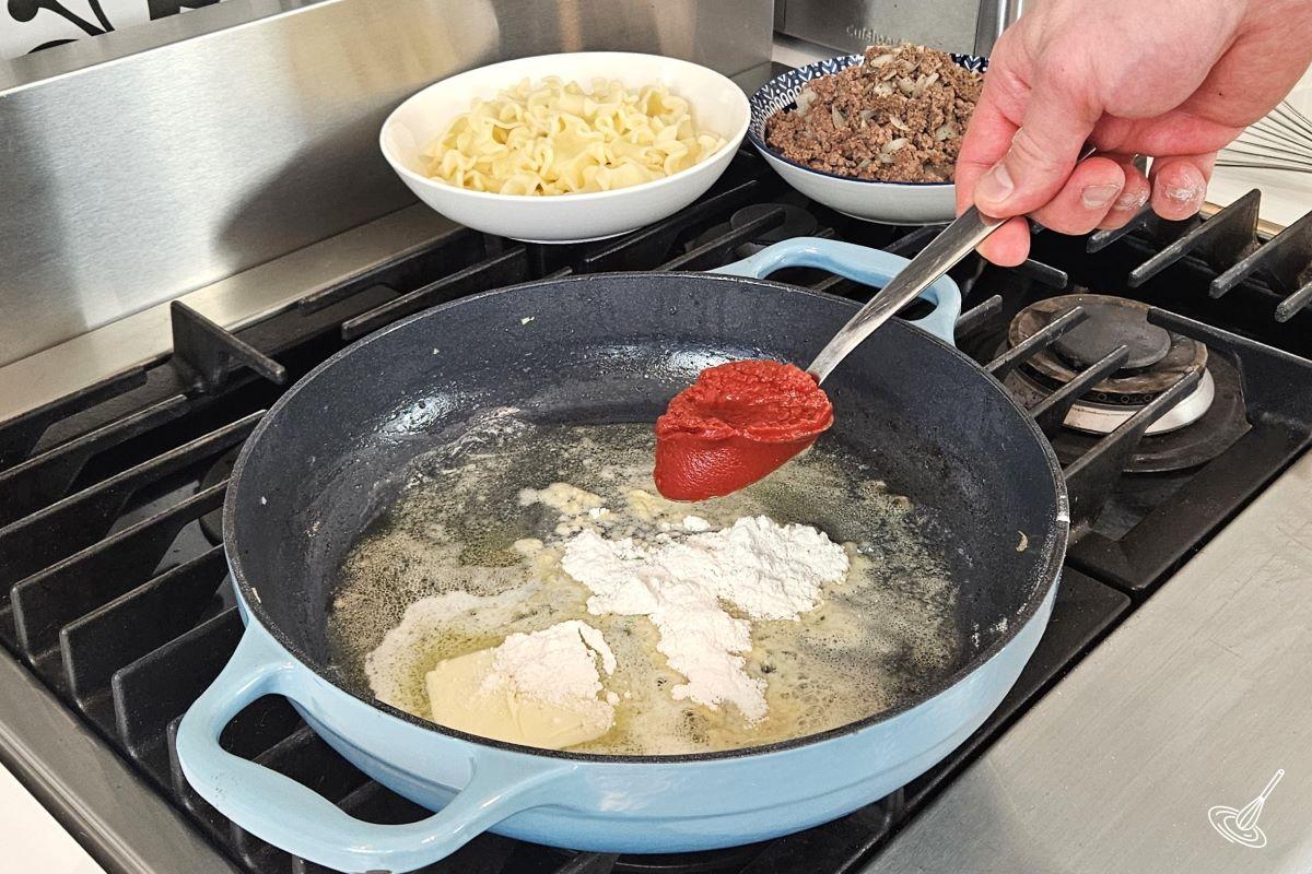 Someone placing tomato paste in a large skillet with melted butter and flour. 