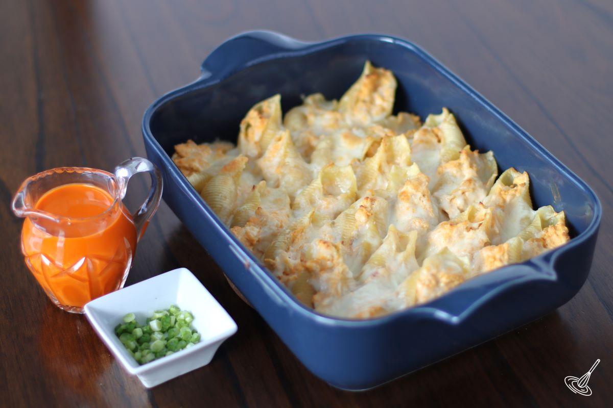 A baking dish of baked Buffalo Chicken Pasta Shells with side dishes of green onion and buffalo sauce. 