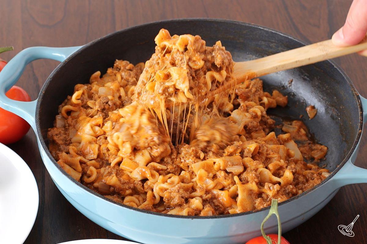 Someone stirring in grated cheese into a skillet of beefaroni recipe.