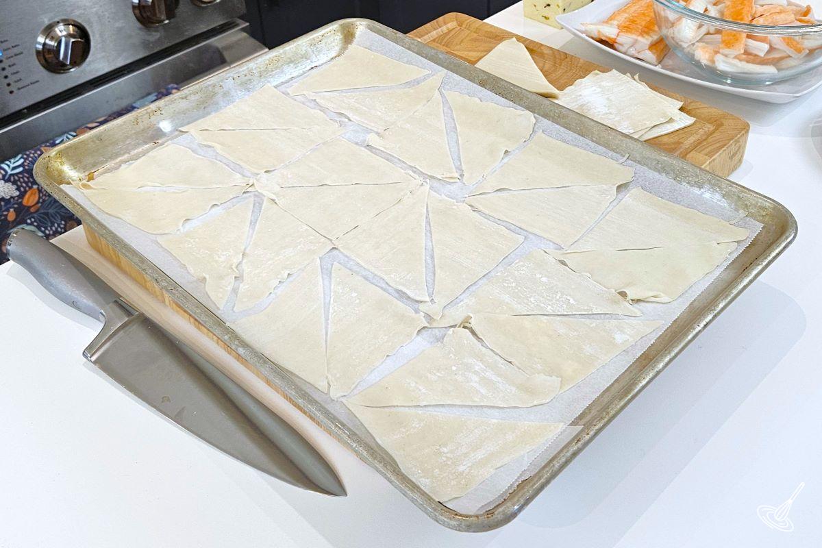 A baking tray with raw wonton wrappers cut in half.