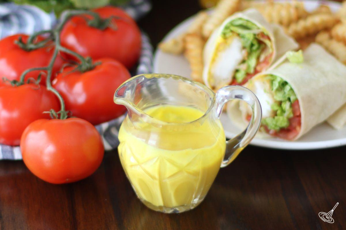 A jar of honey mustard on a table with a Honey Mustard Chicken Wrap in the background. 