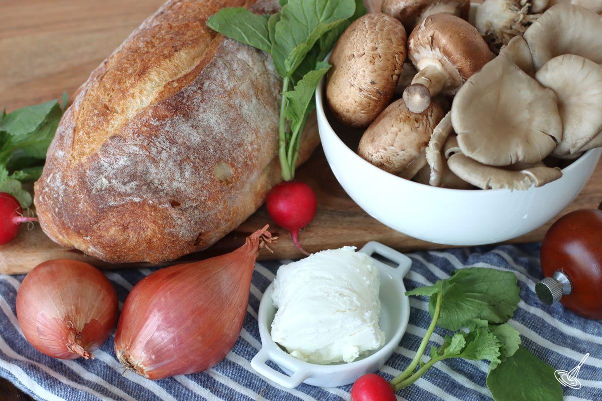 A bread and a bowl of mushroom on a wooden cutting board.