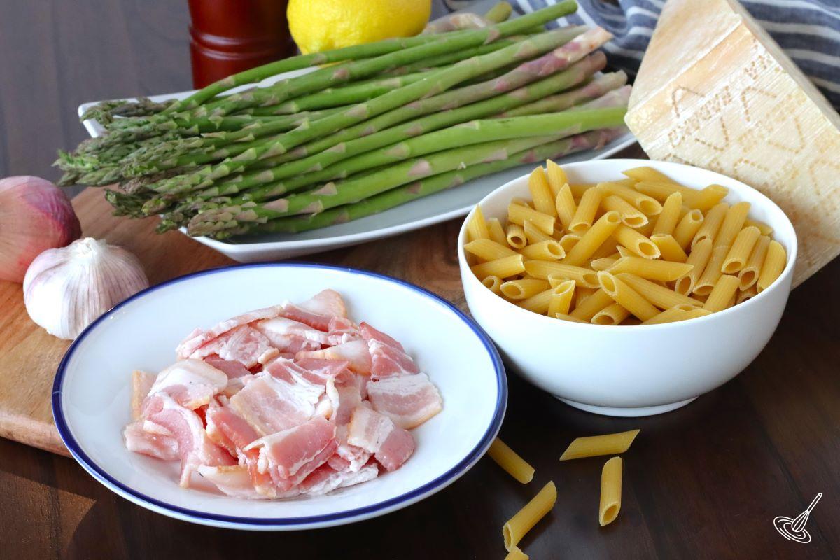 Ingredients on a table including uncooked pasta in a bowl, asparagus on a plate and chopped bacon on a plate.