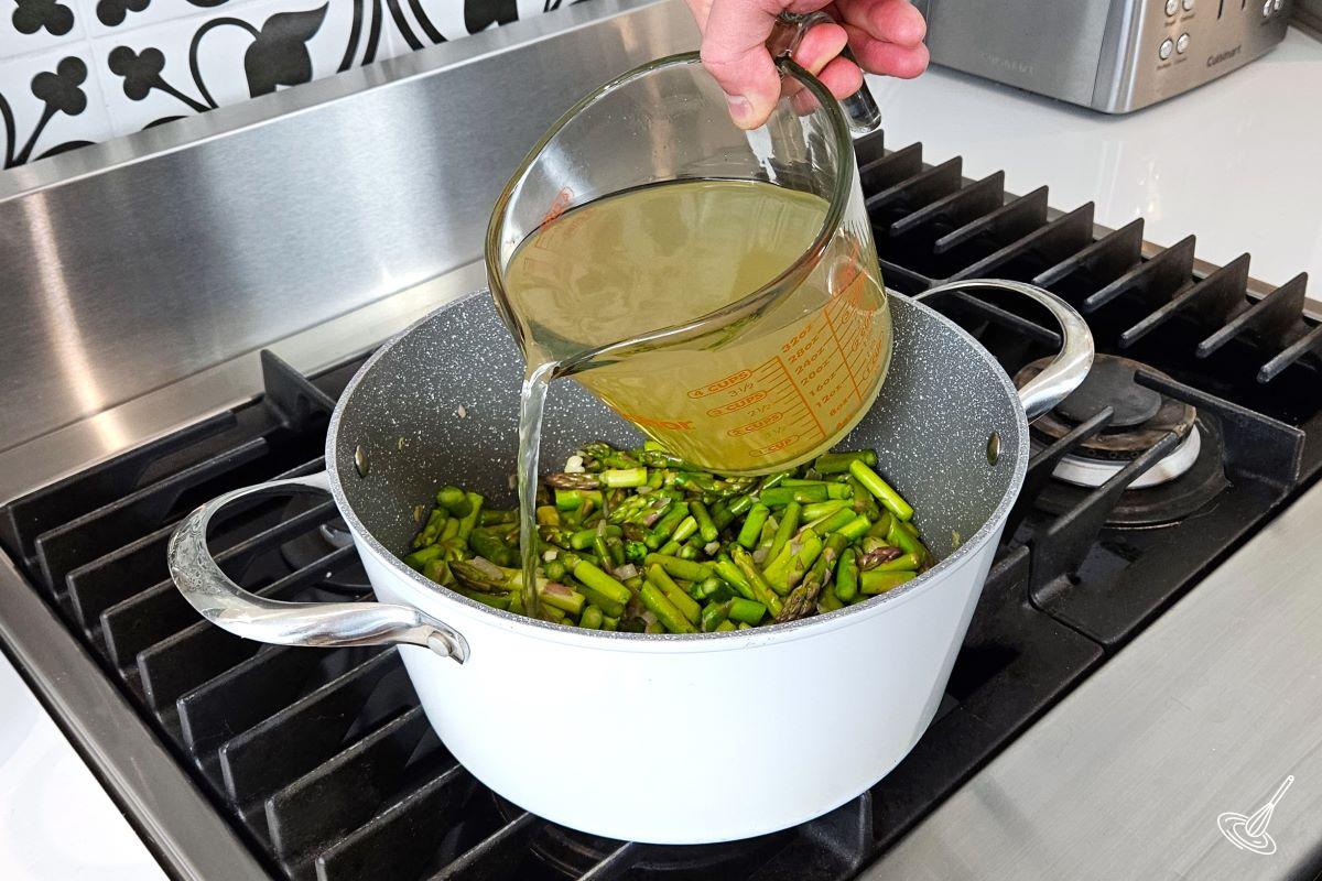 Someone pouring broth into a soup pot containing vegetables. 