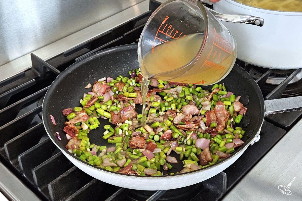 Someone pouring chicken broth into a frying pan containing bacon and veggies.