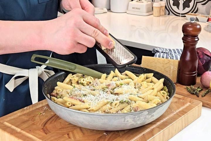 Someone grating parmesan cheese into a Creamy pasta with asparagus and bacon. 