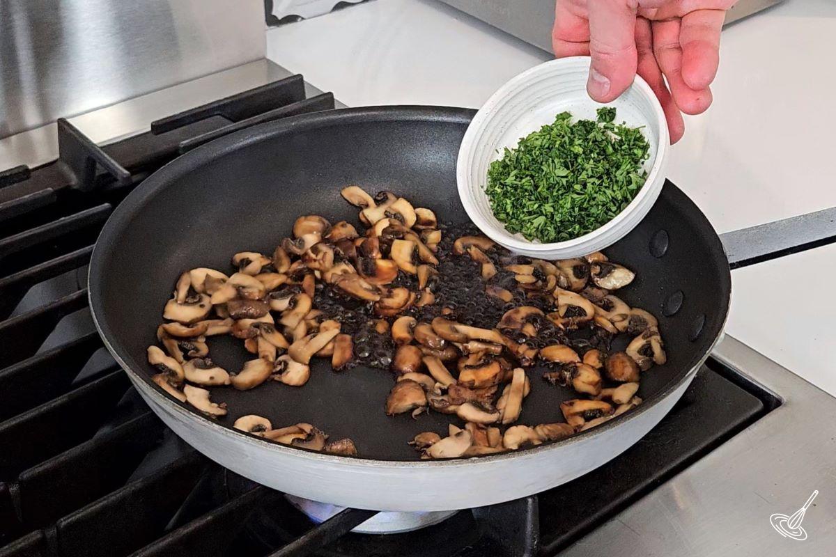 Someone adding chopped parsley to a frying pan with fried mushrooms.