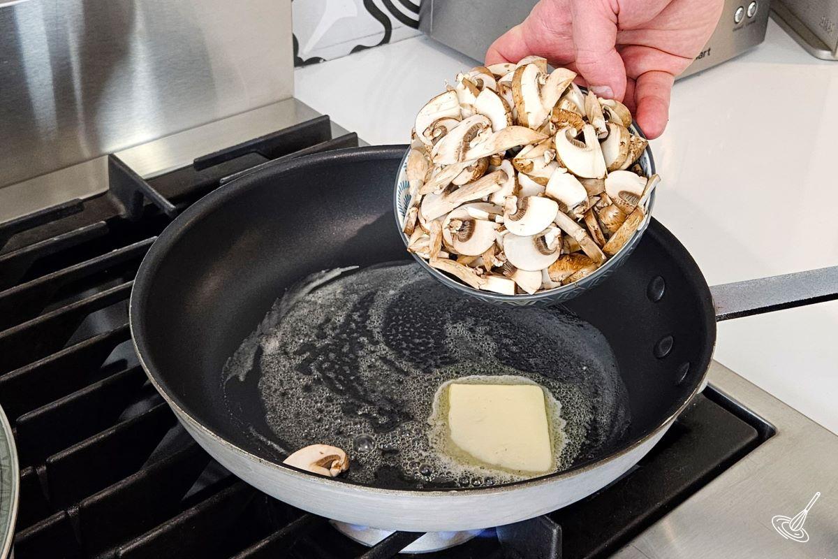 Someone adding sliced mushrooms to a frying pan with melted butter.