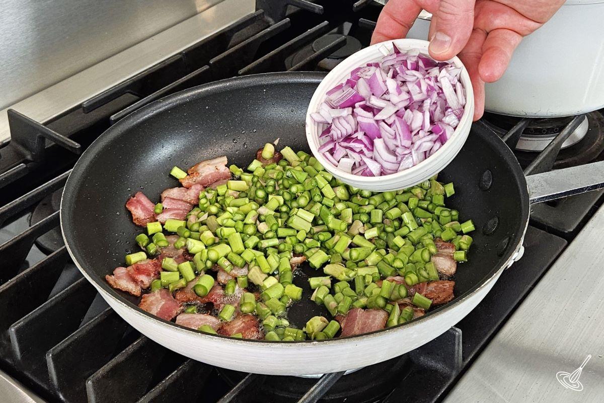 Someone adding red onion to a frying pan containing chopped asparagus and cooked bacon.