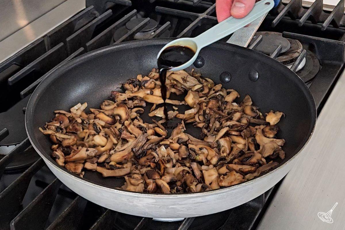 Someone pouring balsamic vinegar in a frying pan with cooked mushrooms and onions. 