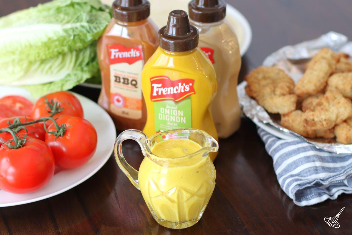 A container of Honey Mustard on a table with a plate of sliced tomatoes and chicken tenders.