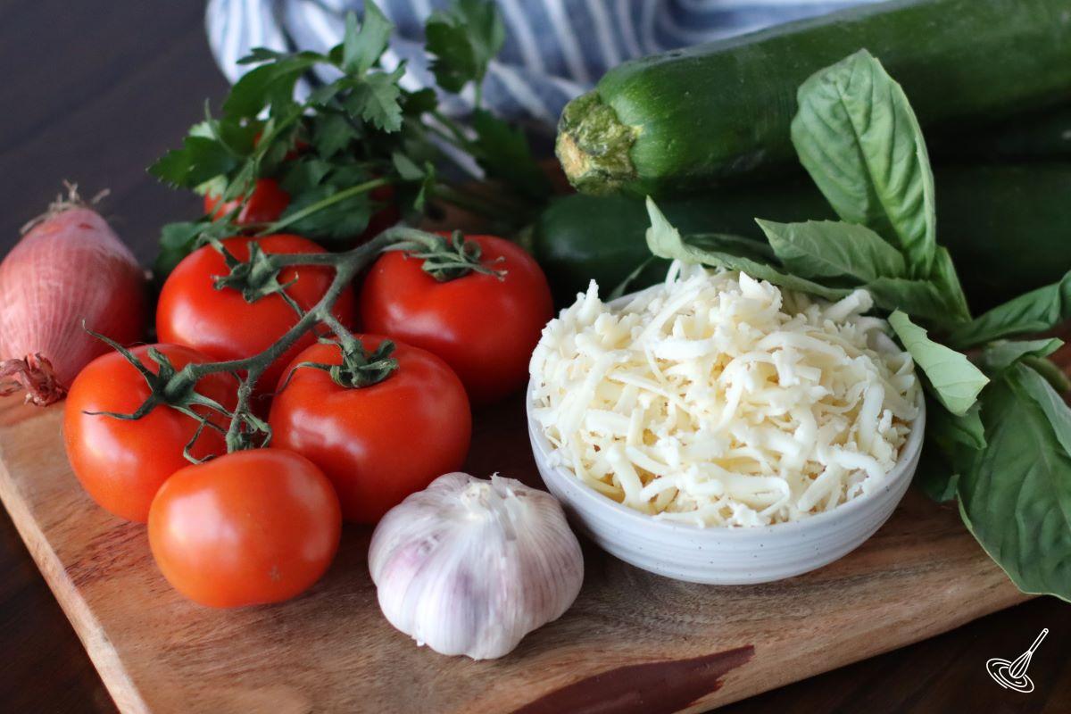 Ingredients on a wooden board including tomatoes, garlic, zucchini and grated mozzarella. 