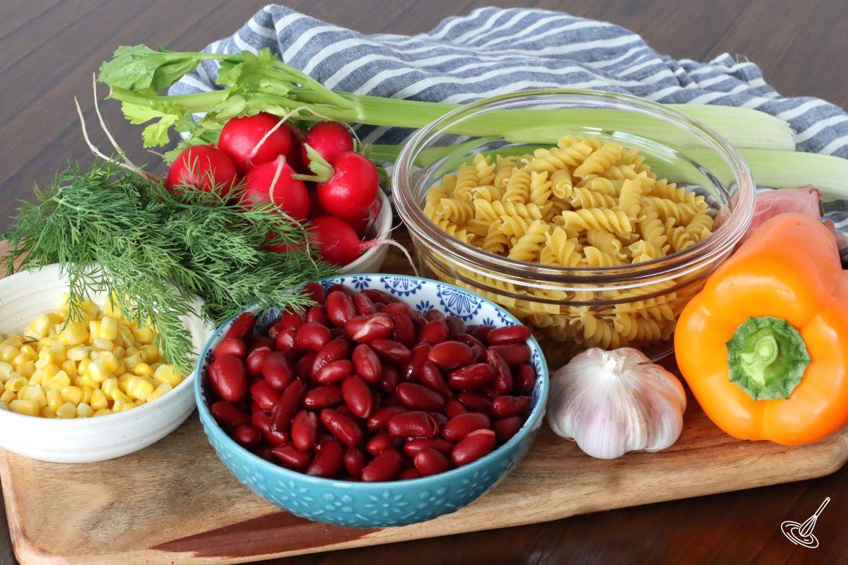 Ingredients on a cutting board, including raw pasta, radishes, corn kernels, garlic, bell pepper and kidney beans.