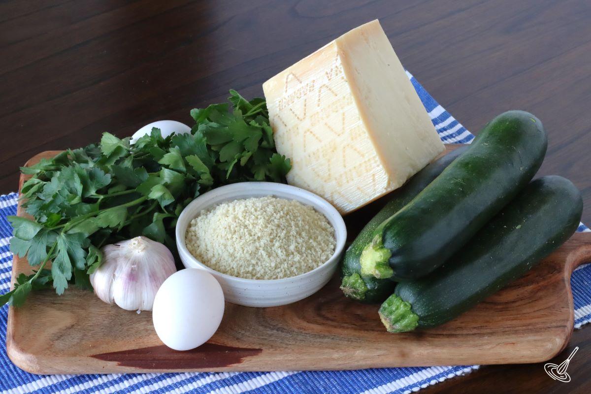 Ingredients on a wooden board including; zucchini, panko crumbs, eggs, parmesan and fresh herbs. 