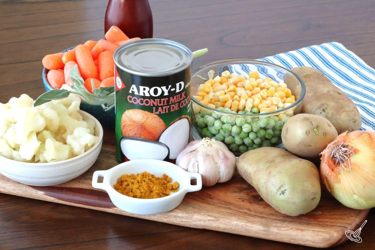 Ingredients on a cutting board, including garlic, potato, onion, curry powder, cauliflower, carrot, coconut milk, peas, and corn. 