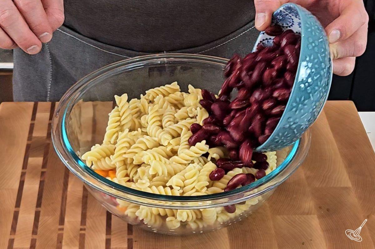 Someone adding kidney beans to a large bowl with cooked pasta and vegetables.