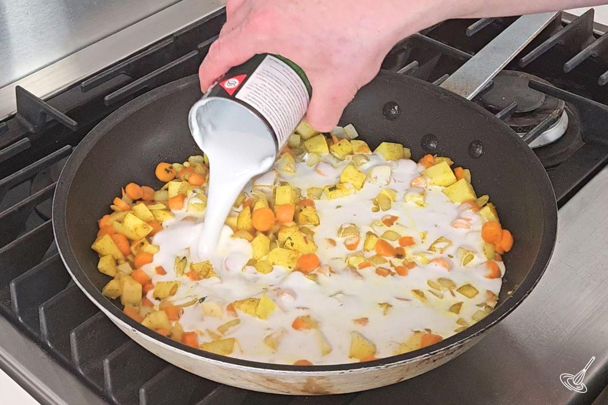 Someone pouring out a can of coconut milk into a large frying pan with curry vegetables.