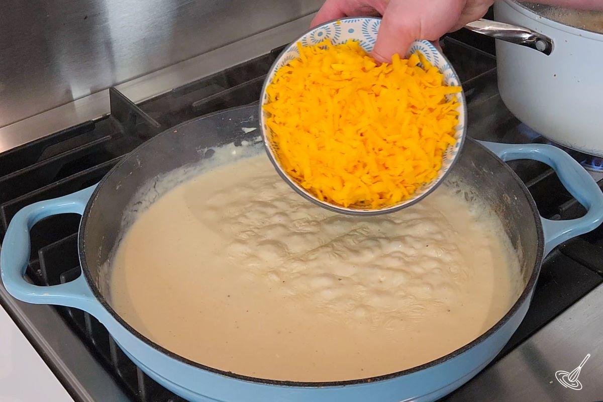 Someone placing grated cheddar cheese in a cream sauce. 