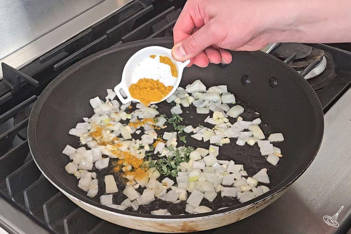 Someone sprinkling curry powder in a large frying pan with onions.