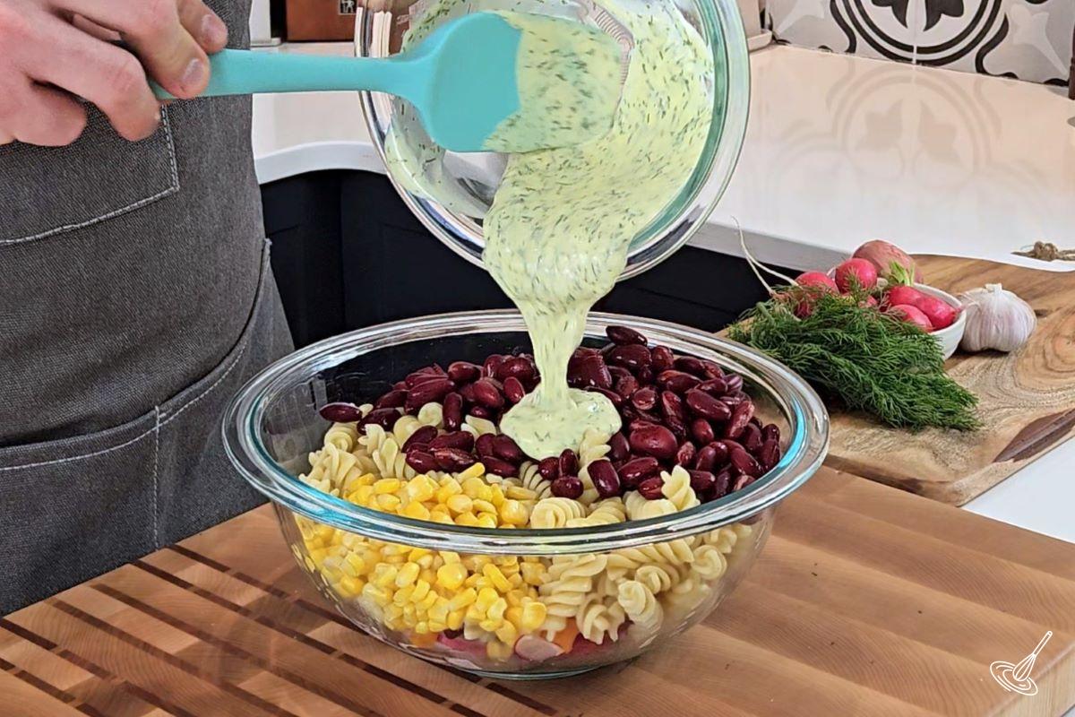 Someone pouring salad dressing in a bowl of pasta salad with kidney beans. 