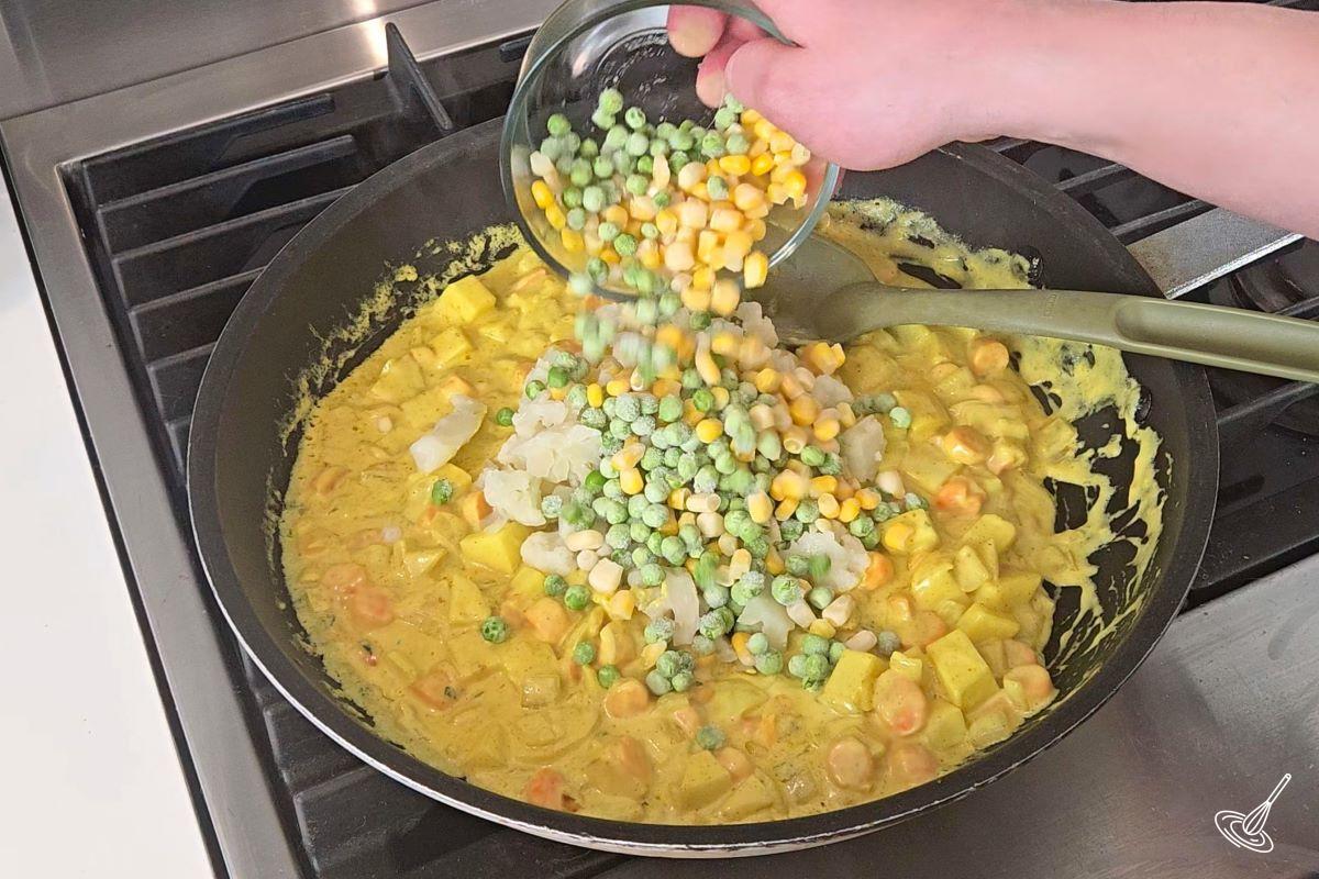 Someone add frozen peas and corn into a large pan with curry vegetable filling.
