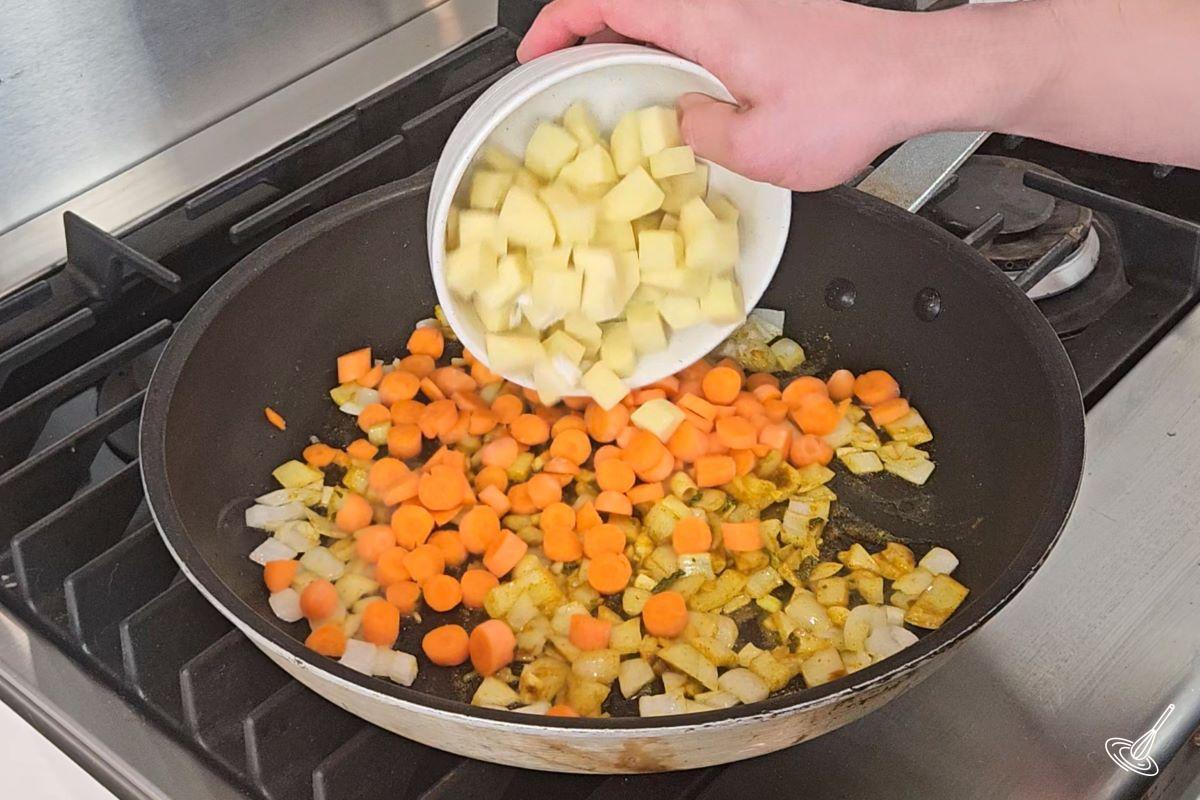Someone placing cubed potatoes in a large frying pan already containing carrot and curry vegetables.