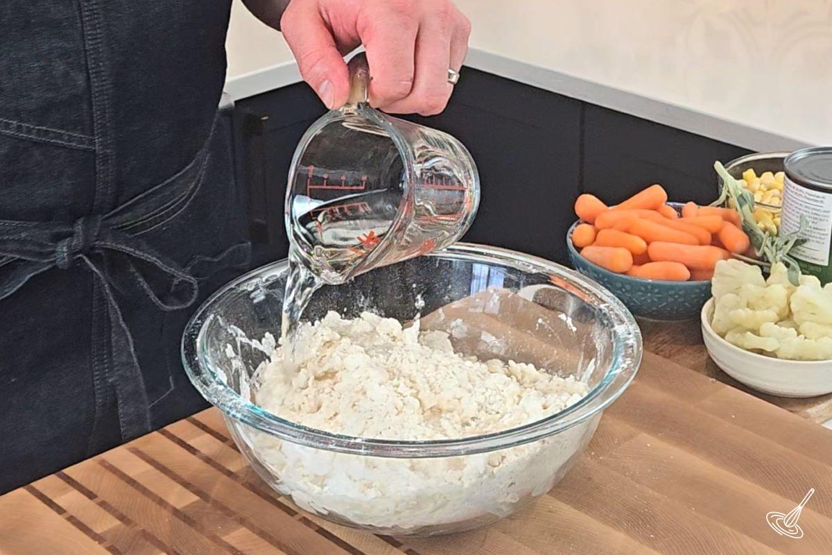 Someone pouring water in a bowl of flour to make pie dough. 