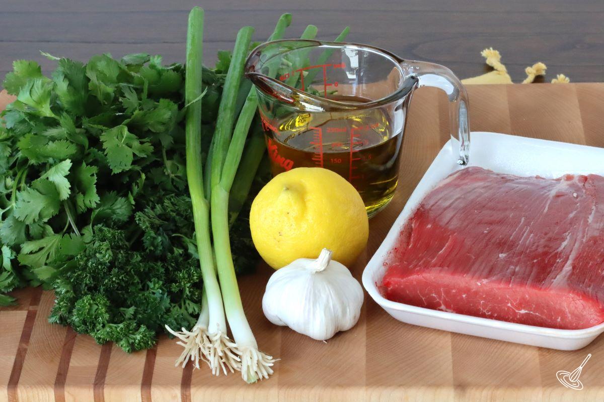 Ingredients on a cutting board including; cilantro, parsley, green onion, lemon, olive oil and flank steak.