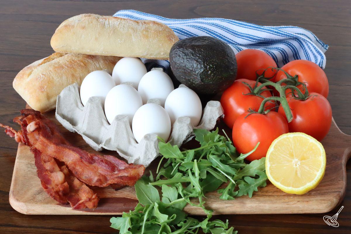 Ingredients on a cutting board including; eggs, tomatoes, avocadoes, bacon, and ciabatta rolls.