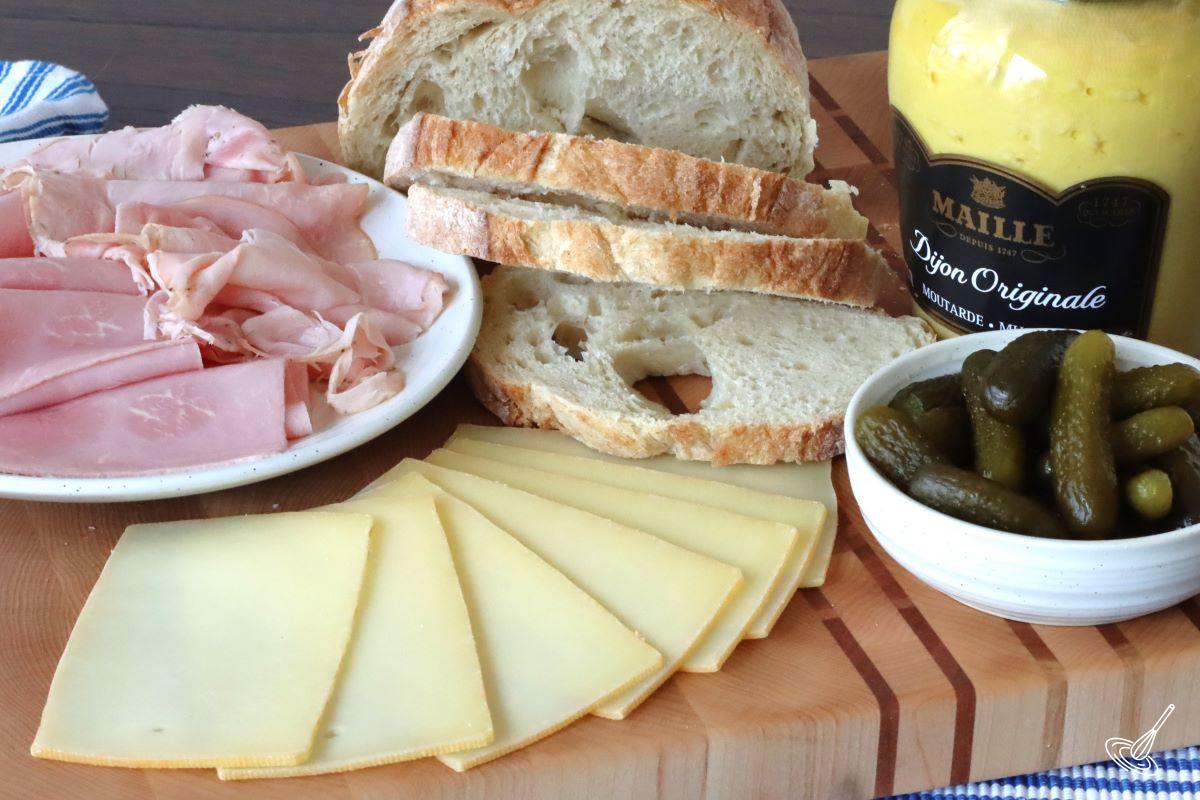 Ingredients on a wooden board, including raclette cheese, sliced ham, gherkin pickles, slices of bread, and mustard.