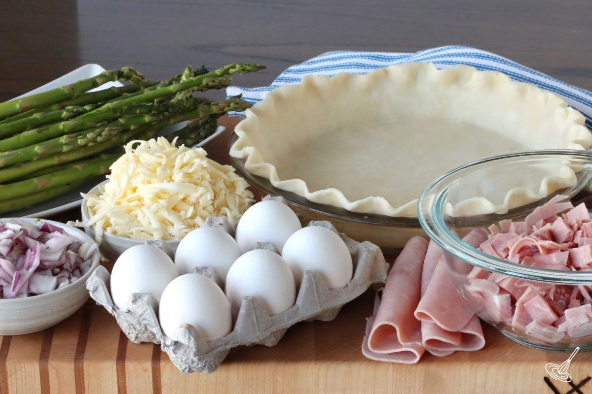 Ingrédients d'une planche à découper, notamment du jambon, des œufs, une pâte à tarte et des asperges. 